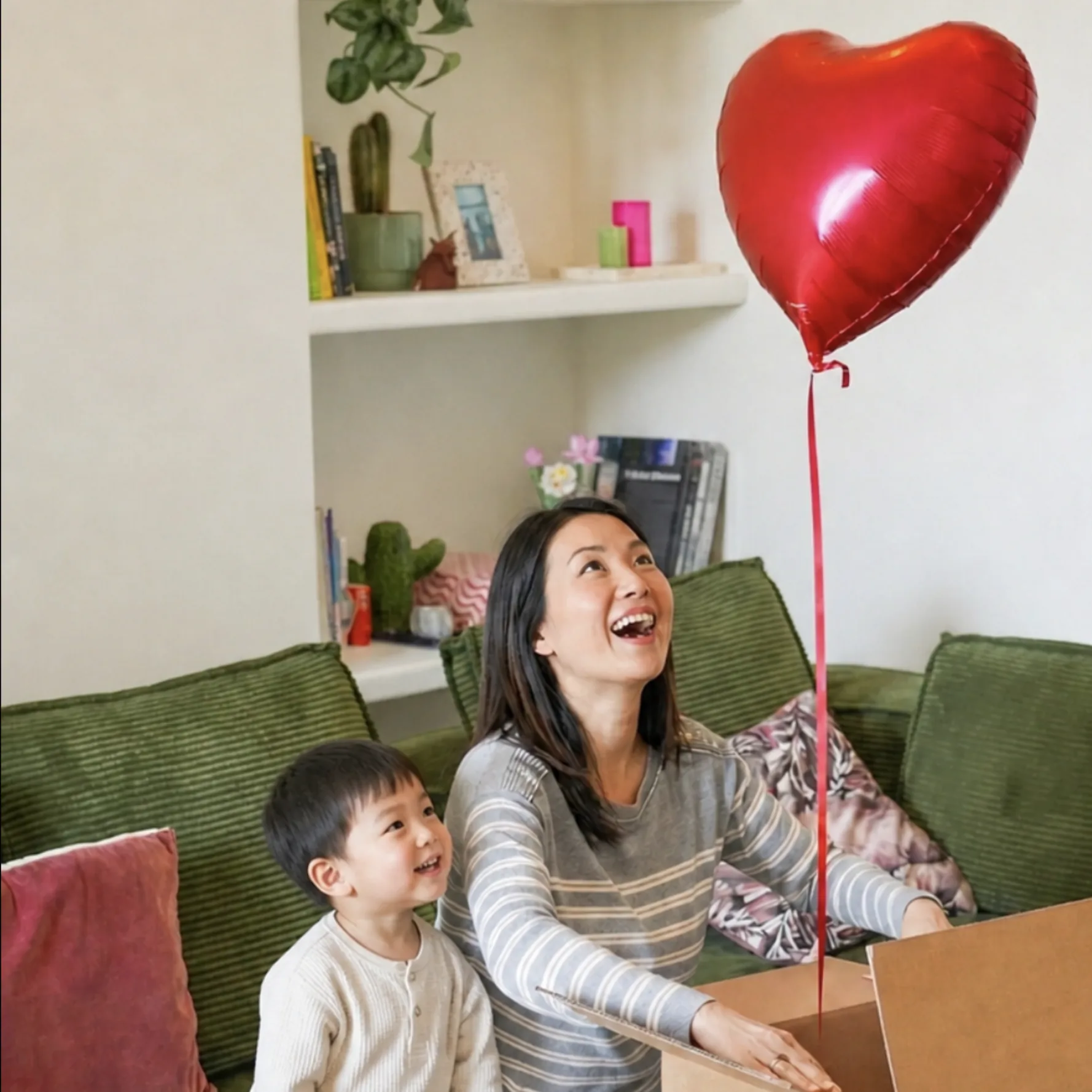A woman and a child on a green sofa opening a box, with a red heart balloon above