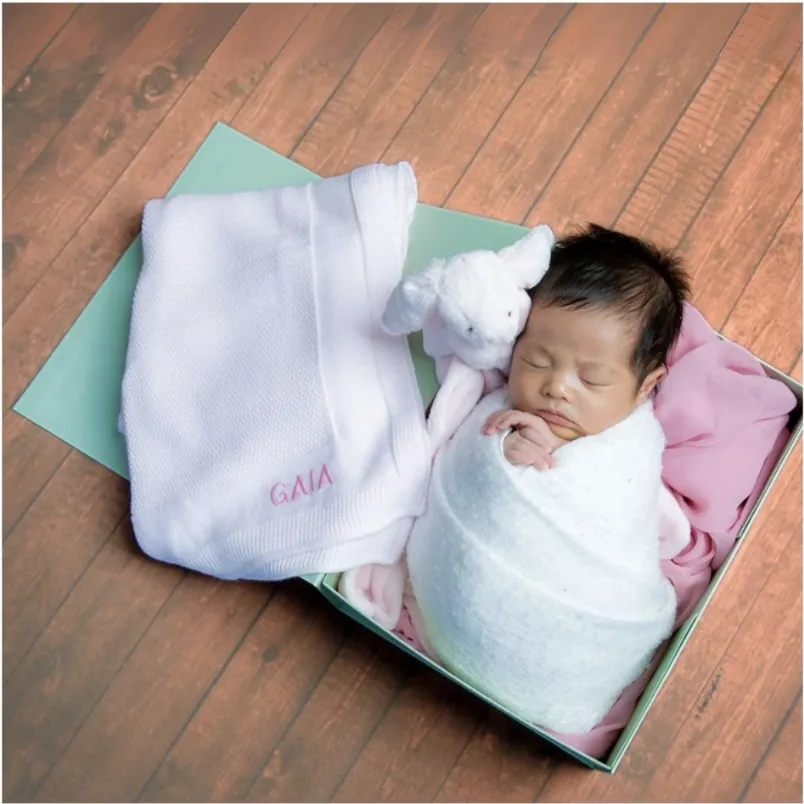 A sleeping baby in a gift box beside a white blanket embroidered with a name and a soft bunny