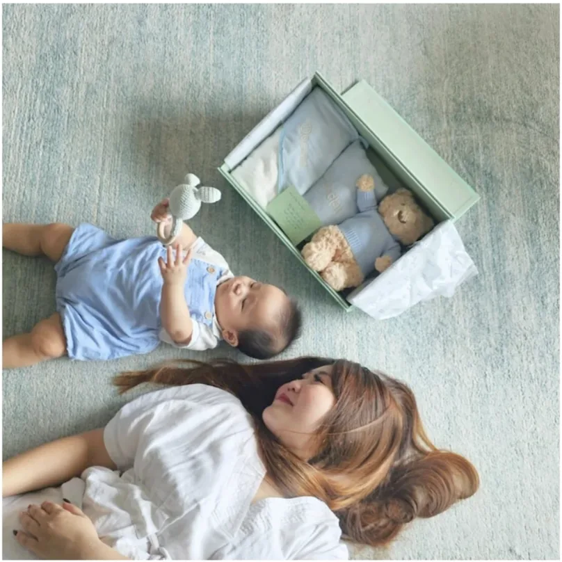 A parent and baby lying beside an open gift box with clothes and a teddy